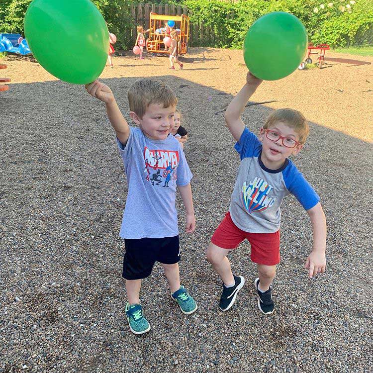 Two boys holding green balloons, one boy is looking at the other as he makes a goofy dance move with his balloon up in the air