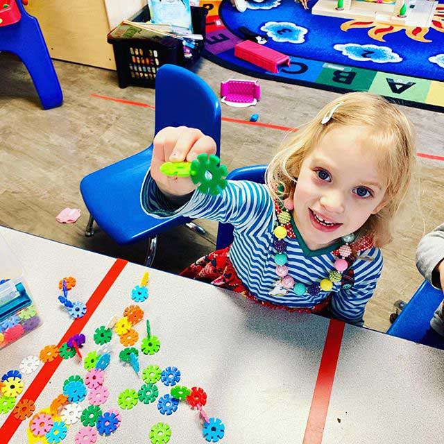 Girl from class holding up a toy, sitting at a table with toys, very happy